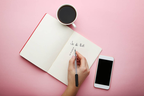 Female hand writing to do list in notepad and cup of coffee on pink background - Фото, изображение