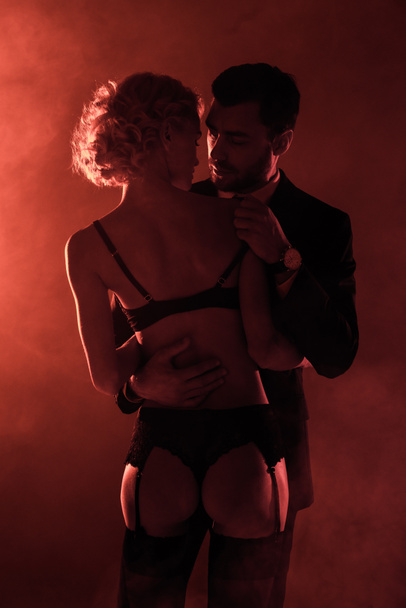 Back view of seductive woman in lingerie looking at man in costume on red smoke background - Foto, afbeelding