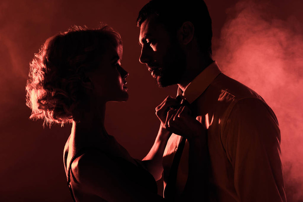 Beautiful couple of young adults looking at each other on red smoke background - Foto, Imagen