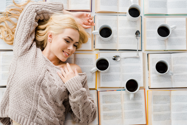 Lachende vrouw leggen in de buurt van klok gemaakt van witte kopjes met koffie en lepels op open boeken - Foto, afbeelding