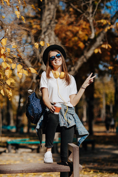Image of a bright smiling hipster girl with brown hair wearing a hat, sunglasses and backpack holding cup of tea or coffee in the park - Φωτογραφία, εικόνα