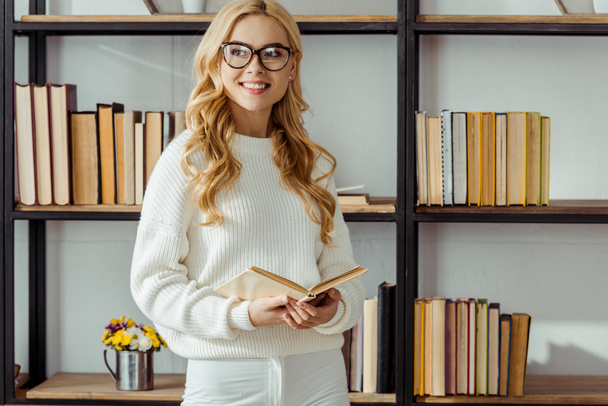 close up of cheerful woman in glasses with book standing near rack - Photo, Image