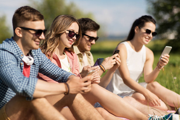 smiling friends with smartphones sitting on grass - Fotó, kép