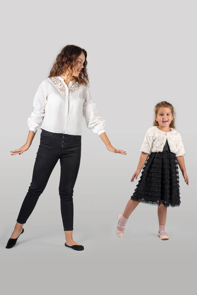 Ολόσωμο πορτρέτο του ένα χαμογελαστό κοριτσάκι φορώντας όμορφο μαύρο κομψό φόρεμα και νεαρή γυναίκα - μητέρα ή αδελφή. Αυτοί είναι ο χορός σε ένα λευκό φόντο στο στούντιο - Φωτογραφία, εικόνα