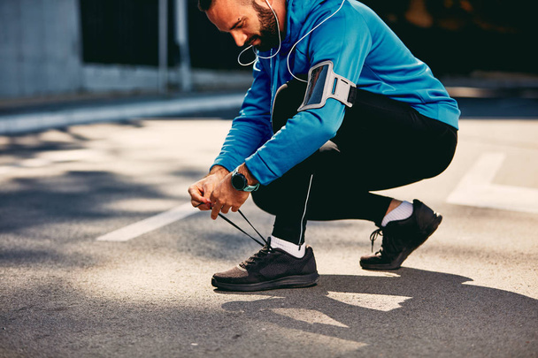 Male runner crouching and tying shoelace. Sportswear on, earphones in ears. Healthy lifestyle concept. - 写真・画像