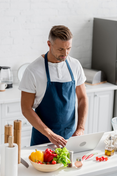 handsome man in apron standing at kitchen and working on laptop - Foto, Bild