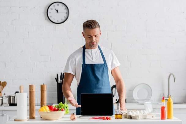 Erwachsener Mann mit schwarzem Bildschirm auf geöffnetem Laptop steht in Küche - Foto, Bild