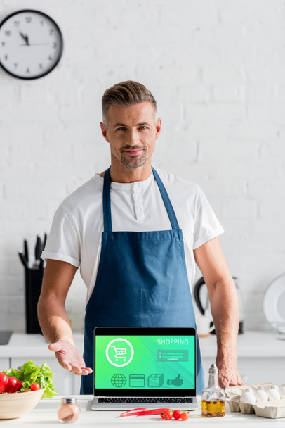 hombre adulto guapo de pie con ilustración de compras en línea en la pantalla del ordenador portátil en la mesa de la cocina
 - Foto, Imagen