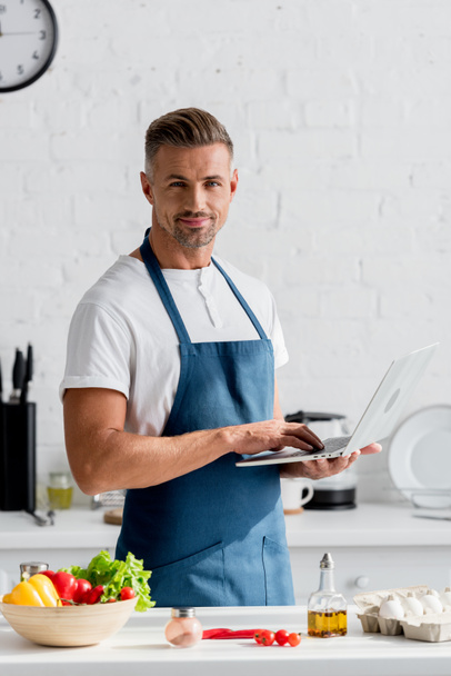 aikuinen mies kannettava tietokone käsissä seisoo keittiössä
 - Valokuva, kuva