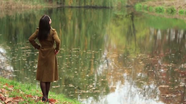 若いスリム女性秋の公園 (1080、25 fps の反射と湖で茶色のコート スタンド weared) - 映像、動画
