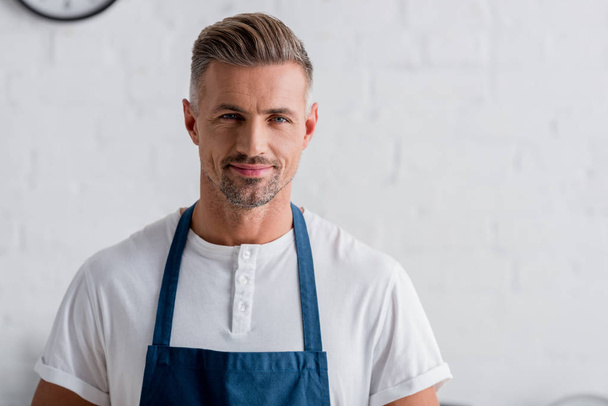 volwassene Glimlachende man in schort staande op keuken - Foto, afbeelding