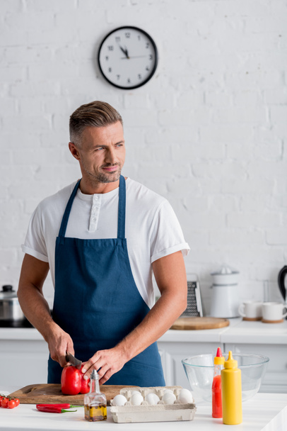 όμορφος ενήλικος άντρας στέκεται στην κουζίνα και την κοπή κόκκινο πιπέρι κουδουνιών - Φωτογραφία, εικόνα
