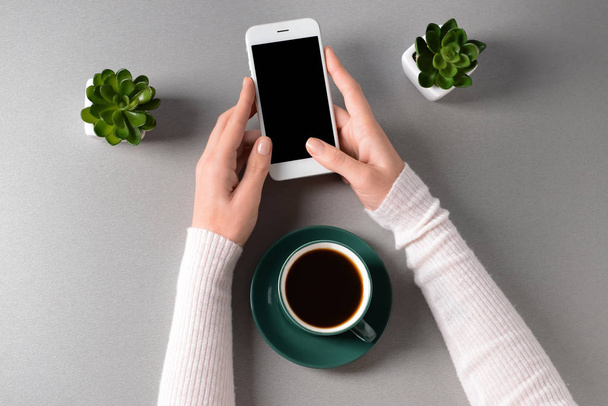 Naisten kädet matkapuhelimella ja kupillinen kahvia kevyellä taustalla, ylhäältä
 - Valokuva, kuva