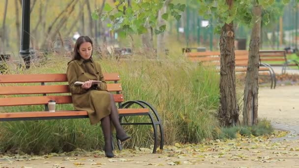 Mujer joven se sienta en un banco y leer el libro en un parque de otoño beber café de una taza de papel (1080p, 25fps, tiro largo
) - Imágenes, Vídeo