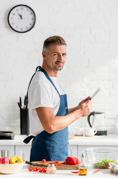 красивый мужчина в фартуке с цифровым планшетом на кухне
 - Фото, изображение