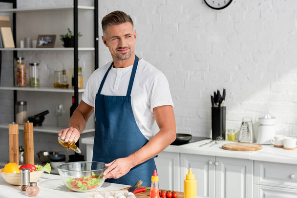 Erwachsener lächelnder Mann fügt Öl in Salat in der Küche hinzu - Foto, Bild