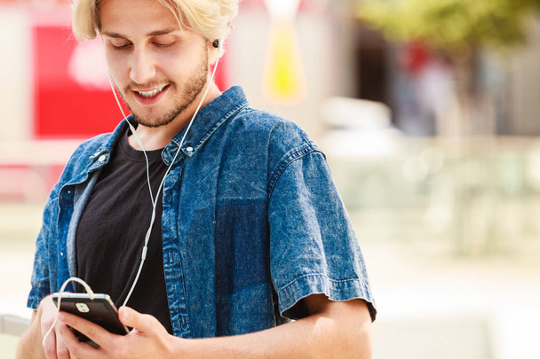 Men fashion, technology, urban style clothing concept. Hipster smiling guy standing on city street wearing jeans outfit listening to music through earphones - Photo, Image