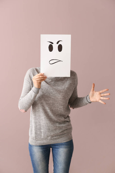 Young woman hiding face behind sheet of paper with drawn emoticon on color background - Photo, Image