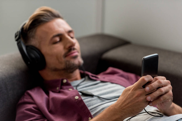 happy man with smartphone and closed eyes listening music while laying on sofa  - Photo, Image