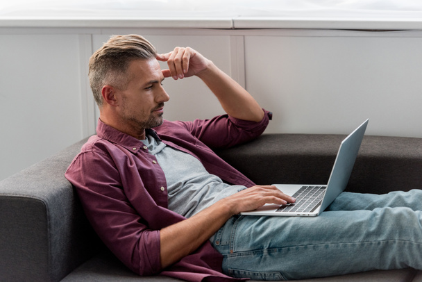 homem pensativo deitado no sofá e trabalhando no laptop no escritório em casa
 - Foto, Imagem