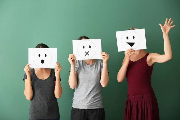 Young women hiding faces behind sheets of paper with drawn emoticons on color background - Photo, Image