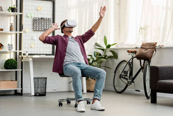 Mann sitzt auf Stuhl und gestikuliert im Virtual-Reality-Headset im Home Office - Foto, Bild