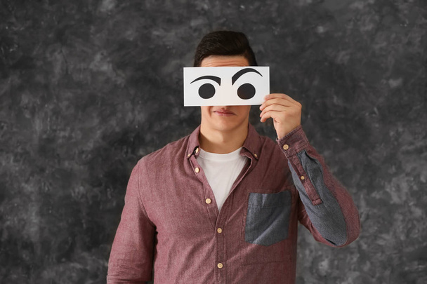 Emotional young man hiding face behind sheet of paper with drawn eyes on grey background - Photo, Image