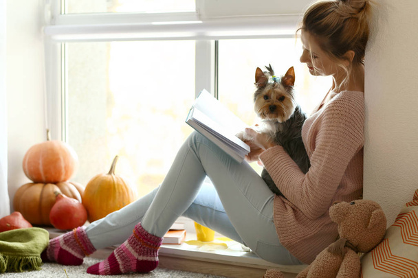 Woman with cute dog reading book at home on autumn day - Foto, Bild