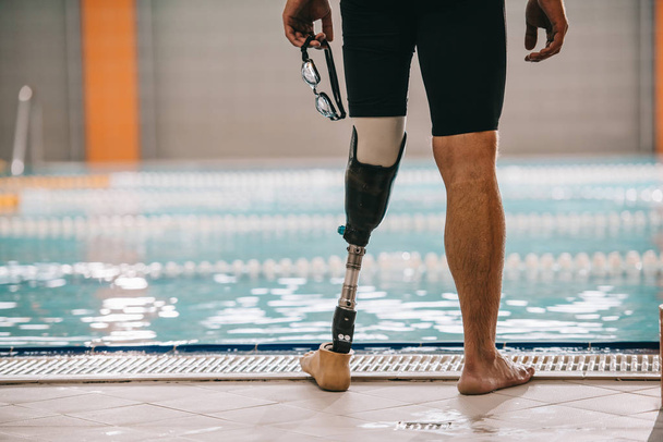 tiro recortado de nadador con pierna artificial de pie frente a la piscina cubierta y la celebración de gafas de natación
 - Foto, imagen