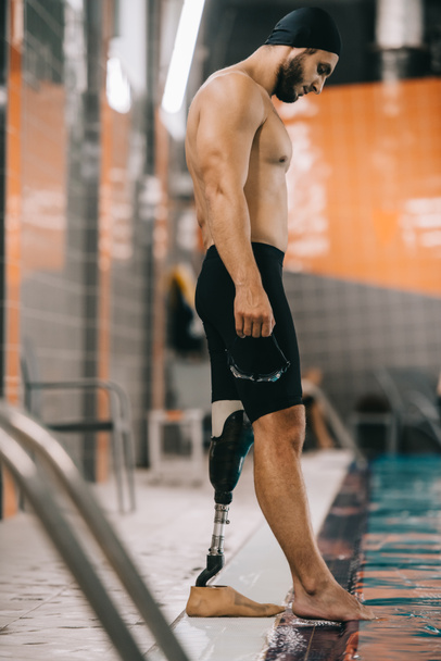 handsome young swimmer with artificial leg standing on poolside at indoor swimming pool and checking water temperature - Zdjęcie, obraz