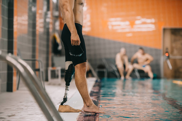 cropped shot of muscular sportsman with artificial leg standing on poolside at indoor swimming pool and checking water temperature - Foto, imagen
