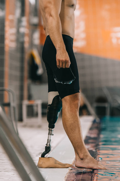 cropped shot of swimmer with artificial leg standing on poolside at indoor swimming pool and checking water temperature - Fotó, kép