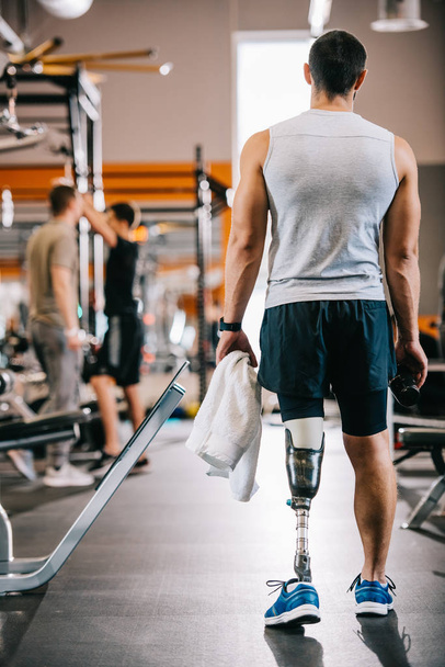 vista trasera del deportista con pierna artificial de pie en el gimnasio y la celebración de la toalla
 - Foto, Imagen