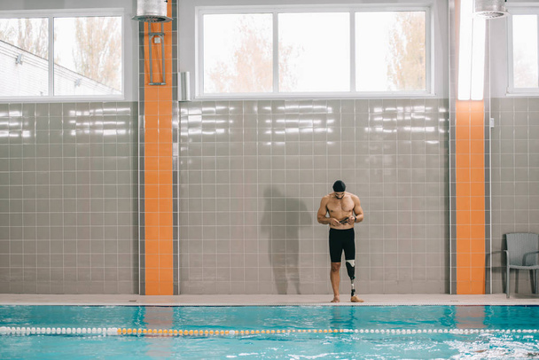 handsome young sportsman with artificial leg standing on poolside at indoor swimming pool - Foto, Bild