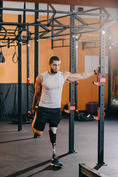 handsome young sportsman with artificial leg stretching near gymnastics ladder at gym - Zdjęcie, obraz