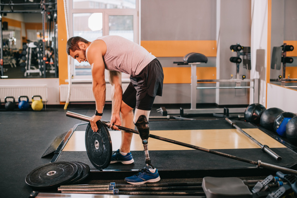 όμορφος νεαρός αθλητής με τεχνητό πόδι βάζοντας στο πιάτο βάρος σε μπαρ στο γυμναστήριο  - Φωτογραφία, εικόνα