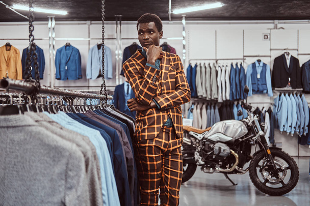 Elegantly dressed African man posing with hand on chin while standing in a classic menswear store. - Foto, Bild