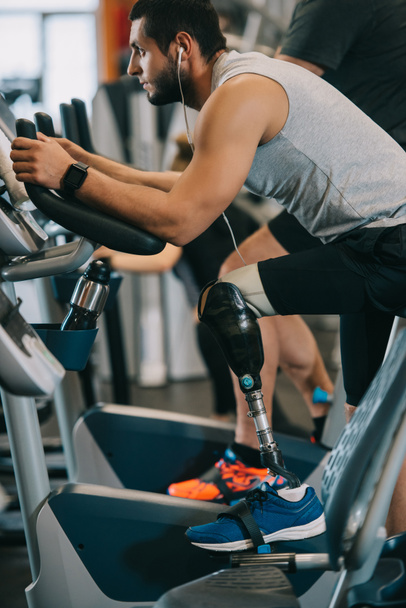 Seitenansicht eines hübschen jungen Sportlers mit Beinprothese, der auf einem stationären Fahrrad im Fitnessstudio trainiert - Foto, Bild