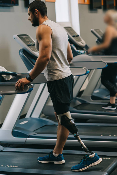 handsome young sportsman with artificial leg walking on treadmilsl at gym - Photo, Image