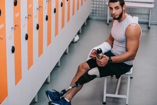 handsome young sportsman with artificial leg sitting on bench at gym changing room and looking at camera - Foto, imagen