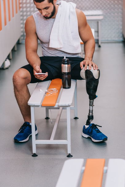 attractive young sportsman with artificial leg sitting on bench at gym changing room and using smartphone - Fotografie, Obrázek