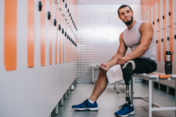 usměvavý mladý sportovec s umělou nohou sedí na lavičce v tělocvičně, šatnou a pomocí smartphone - Fotografie, Obrázek
