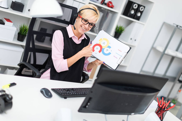 ein junges Mädchen mit Kopfhörern sitzt an einem Computertisch und hält ein Blatt in einem Diagramm. - Foto, Bild