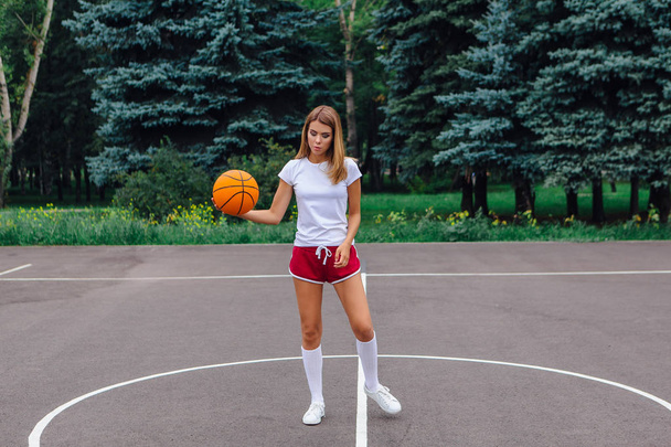 Beautiful young blonde girl dressed in white t-shirt, shorts and sneakers, plays with ball on a basketball court outdoors. Copy space. - 写真・画像