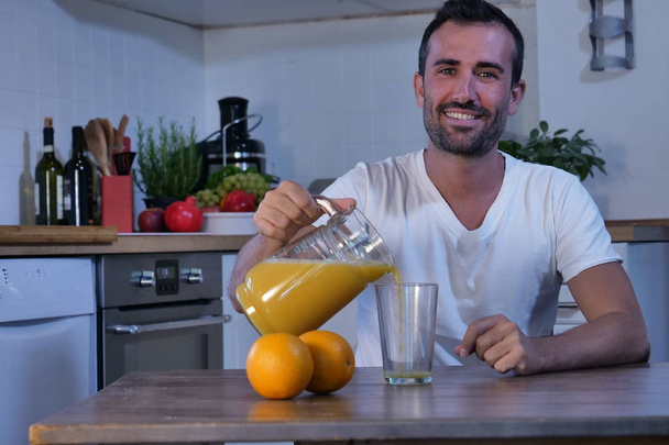 smiling Caucasian man sitting at kitchen table and poring squeezed orange juice from jar into glass and looking at camera - Photo, Image