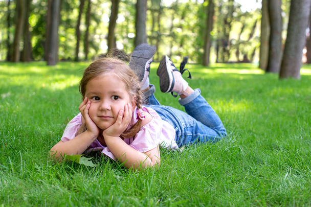 La niña está tumbada en la hierba verde del parque y mira soñadoramente a la distancia.
 - Foto, Imagen