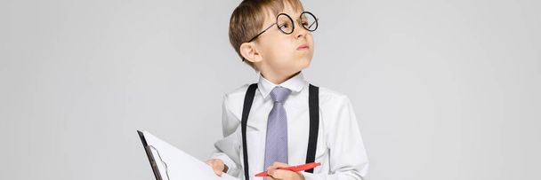 Beyaz bir gömlek, pantolon askısı, kravat ve ışık kot büyüleyici bir çocuk gri bir arka plan üzerinde duruyor. Çocuk tutan bir kalem ve notlar için yaprak - Fotoğraf, Görsel