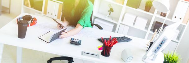 Une jeune fille avec des lunettes dans le bureau s'assoit à la table et tient un journal intime et un téléphone dans ses mains
. - Photo, image