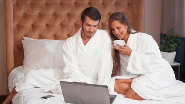 junges Paar beim Kaffee im Bett, während es Laptop benutzt. Die schöne Frau lacht, während sie mit ihrem Mann Kaffee im Bett trinkt. schöner, fröhlicher Mann und seine Frau benutzen gemeinsam Laptop im Hotelzimmer - Filmmaterial, Video