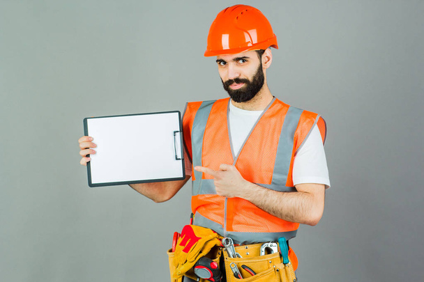 Ein Bauarbeiter mit orangefarbenem Helm auf grauem Hintergrund signalisiert etwas. - Foto, Bild